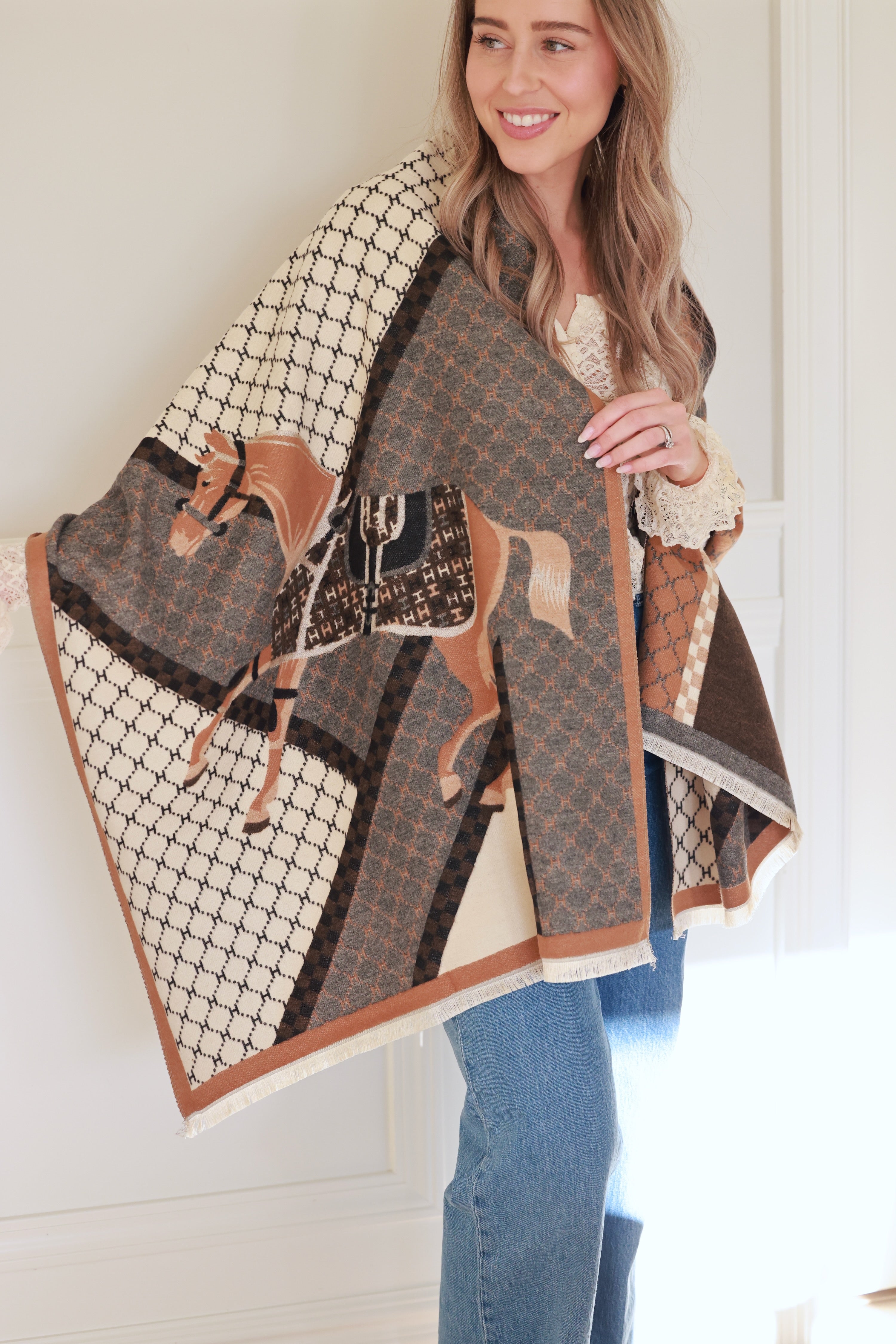 Camel-colored equestrian-inspired scarf with soft cashmere-like texture and subtle horse motif, featuring eyelash fringe and finished hems, styled as a chic wrap for day or evening wear.