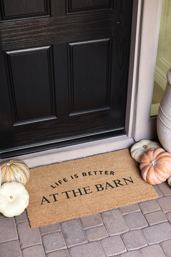 Doormat with "Life is better at the barn" text lies on brick porch. Surrounded by white and striped pumpkins, creating a cozy autumn feel.